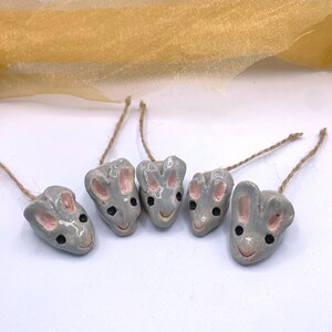 May include: Five grey ceramic bunny ornaments with pink inner ears and black eyes, each with a jute string for hanging. The ornaments are arranged in a row against a white background, with a gold fabric in the background.