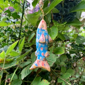 May include: A ceramic fish ornament with a blue body, red spots, and gold accents. The fish is suspended by a gold wire and twine, hanging among green leaves. The fish's fins and tail have a textured, colourful design.