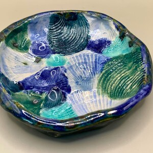 May include: A small, blue and green ceramic dish with a scalloped edge. The dish is decorated with a pattern of seashells.