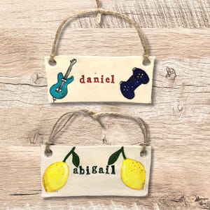 May include: Two rectangular ceramic plaques with twine hangers. The top plaque has a blue guitar and a black game controller, with the name "daniel" in red. The bottom plaque features two yellow lemons and the name "abigail" in green.
