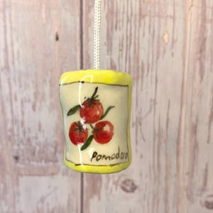 May include: A cylindrical ceramic light pull featuring a hand-painted design of three red tomatoes with green leaves on a cream background, framed by a bright yellow border. The word Pomodoro is written in black script at the bottom.