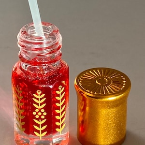 May include: A small, clear glass bottle with a gold lid and a red liquid inside. The bottle has a gold design of leaves and flowers on the side. The bottle comes with a white dropper.