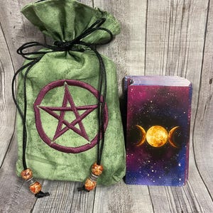May include: A green velvet drawstring bag with a purple pentagram design and a deck of tarot cards. The tarot cards have a cosmic design with a full moon and crescent moons. The bag has decorative beads on the drawstrings.