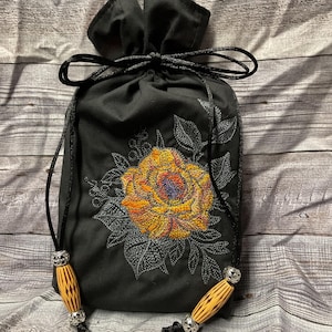 May include: Black drawstring bag with a yellow embroidered rose and green leaves. The bag has wooden beads with silver accents on the drawstrings.