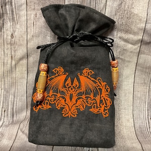 May include: A dark gray drawstring bag with an embroidered orange bat and ornate design. The bag has a black drawstring closure and decorative orange and brown beads on the drawstrings. The bag is made of a soft fabric.