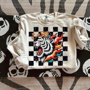 May include: A cream colored sweatshirt with a black and white checkered pattern. The pattern features a white tiger with orange and blue flames. The word "easy" is written in black on the checkered pattern.