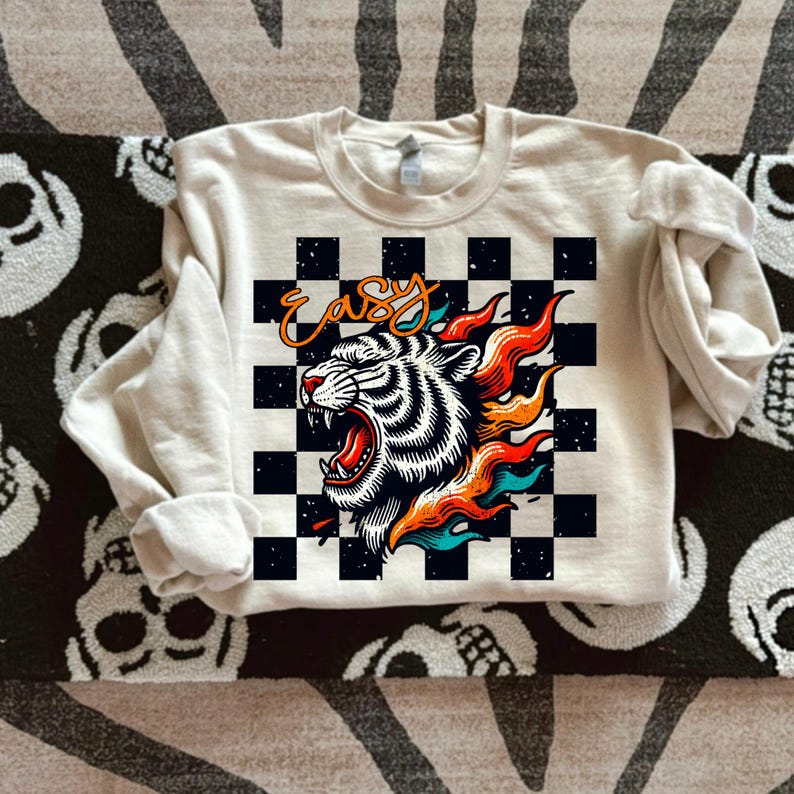 May include: A cream colored sweatshirt with a black and white checkered pattern. The pattern features a white tiger with orange and blue flames. The word "easy" is written in black on the checkered pattern.