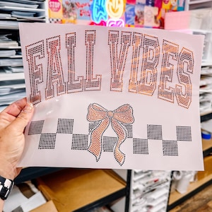 May include: A light pink fabric with the words "FALL VIBES" in a dotted, copper-colored font. Below the text is a copper-colored bow with black and white checkered accents. The fabric is held by a person.