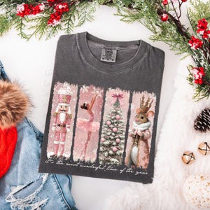 May include: A charcoal gray t-shirt featuring a festive design with a pink nutcracker, ballerina, Christmas tree, and mouse. The shirt has the text "It's the most wonderful time of the year" at the bottom. The shirt is a Comfort Colors brand.