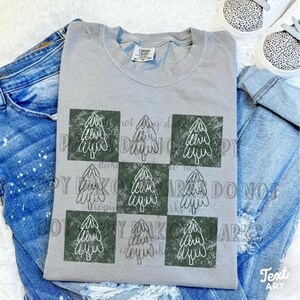 May include: A gray t-shirt featuring a grid pattern of Christmas trees in dark green squares. The shirt is paired with blue jeans and leopard print sneakers. The shirt has a Comfort Colors tag.
