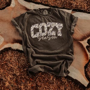 Pode incluir: Uma camiseta cinza escuro com mangas enroladas. A camisa apresenta as palavras "COZY Season" em um padrão de camuflagem e escrita. A camisa é exibida em uma superfície marrom e bege.