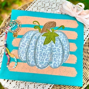 May include: A turquoise shirt featuring a central pumpkin graphic with a light blue leopard print pattern, a brown stem, and a green leaf. The design is layered over horizontal peach-colored brushstrokes. A multi-colored beaded bracelet is attached.
