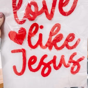 Puede incluir: Tela blanca con el texto rojo "Love like Jesus" y un corazón rojo. El texto está en una fuente de escritura y parece estar hecho de un material texturizado. La tela está parcialmente oscurecida por una lámina de plástico transparente.
