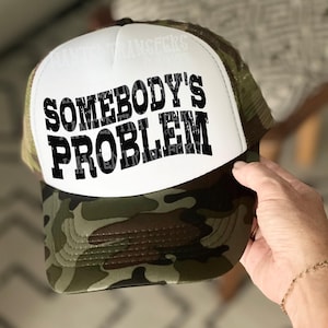 May include: A white and camouflage trucker hat with the text "Somebody's Problem" in black lettering.