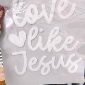 May include: White, fluffy, embroidered patches spelling out "love like Jesus" with a heart symbol. The patches are packaged in a clear plastic bag, ready for application on fabric or other surfaces.
