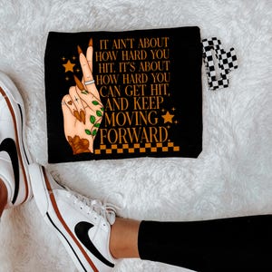 May include: Black makeup bag with a white hand with orange nail polish and a green leaf design. The bag has the text "IT AIN'T ABOUT HOW HARD YOU HIT. IT'S ABOUT HOW HARD YOU CAN GET HIT, AND KEEP MOVING FORWARD."