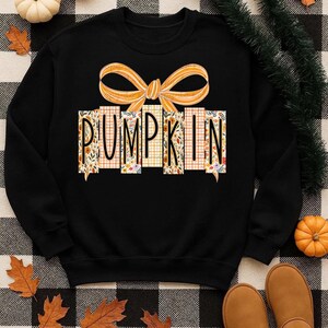 May include: Black sweatshirt with the word "PUMPKIN" in a decorative font. The letters are made of floral and plaid patterns. An orange bow is above the word. The sweatshirt is a fall-themed item.