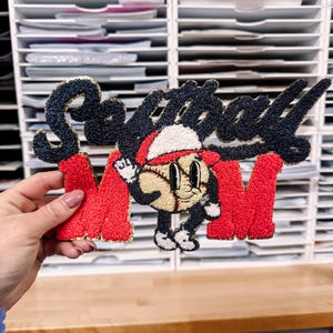 May include: A decorative patch with the words "Softball Mom" in black script and red block letters. A baseball character wearing a Santa hat is in the center. The patch has a textured, embroidered appearance.
