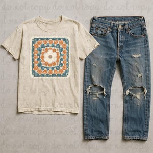 May include: A cream-colored t-shirt with a square floral design in orange, teal, and cream. The shirt is paired with distressed blue denim jeans. The jeans have ripped knees.