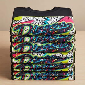 May include: A stack of black t-shirts with a colorful graphic design. The design features the word "Dinger" in a stylized font with a retro, neon aesthetic. The shirts are neatly folded, showcasing the vibrant print.