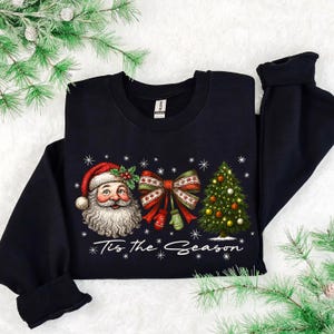 Puede incluir: Sudadera negra con un diseño navideño. El gráfico incluye a Papá Noel, un gran lazo con detalles rojos y verdes, un árbol de Navidad decorado y el texto "Tis the Season" en blanco.