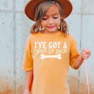 Puede incluir: Una persona joven lleva una camiseta de color amarillo mostaza con la frase "I'VE GOT A bone to pick" en escritura blanca y un gráfico de hueso. La persona también lleva un sombrero marrón.