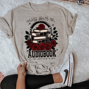 May include: Beige t-shirt with a graphic of a stack of books with headphones and roses. The text reads "unless you're an audiobook, i'm not listening to you."