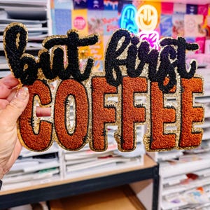 May include: A decorative sign with the phrase "but first COFFEE" in a bold, textured design. The letters are outlined in gold glitter, with black and orange text. The sign is held in front of a shelf.