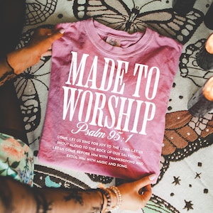 May include: A dusty rose-colored t-shirt with the words "MADE TO WORSHIP" in large white letters. Below, "Psalm 95:1" is printed, followed by a verse. The shirt is folded and held by hands.