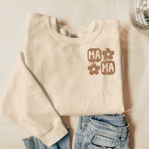 May include: A white sweatshirt with a brown graphic design that says "MA*MA" with a flower between the words.