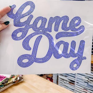 May include: A white sheet with the words "Game Day" spelled out in blue rhinestones. The script is in a cursive font. The sheet is being held by a hand with dark green nail polish.