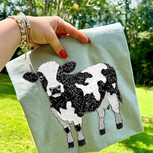 Könnte beinhalten: Ein hellgrünes quadratisches Textil mit einem detaillierten schwarz-weißen Kuhdesign. Die Kuh ist mit kleinen, funkelnden Akzenten verziert. Das Textil wird vor einem verschwommenen grünen Hintergrund gehalten.