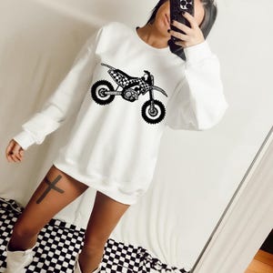 Puede incluir: Sudadera blanca con un diseño de moto de motocross en blanco y negro. La sudadera tiene cuello redondo y mangas largas. El gráfico de la moto presenta un patrón de bandera a cuadros en el asiento y una ilustración detallada del motor y las ruedas.
