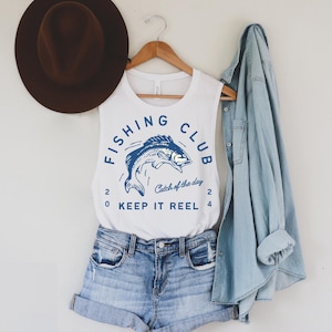 May include: White sleeveless tank top with a blue graphic of a fish jumping out of the water. The graphic says "Fishing Club" with the words "Catch of the day" and "Keep it Reel" underneath.