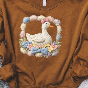 May include: A brown sweatshirt featuring a detailed appliqué of a white duck with blue polka dots, a pink bow, and an orange beak. The duck is surrounded by a floral frame with a blue bow at the bottom. The design is centered on the front of the sweatshirt.