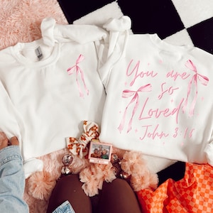 May include: Two white sweatshirts with pink ribbon designs. One sweatshirt has the text "You are so Loved! John 3:16" in pink. The sweatshirts are on a pink surface with decorative items, including a photo frame and a floral hair accessory.
