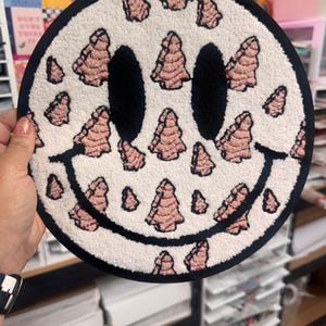 May include: A round, white rug with a smiley face design. The eyes and mouth are black, and the face is surrounded by a black border. The rug is decorated with pink tree-shaped patterns.