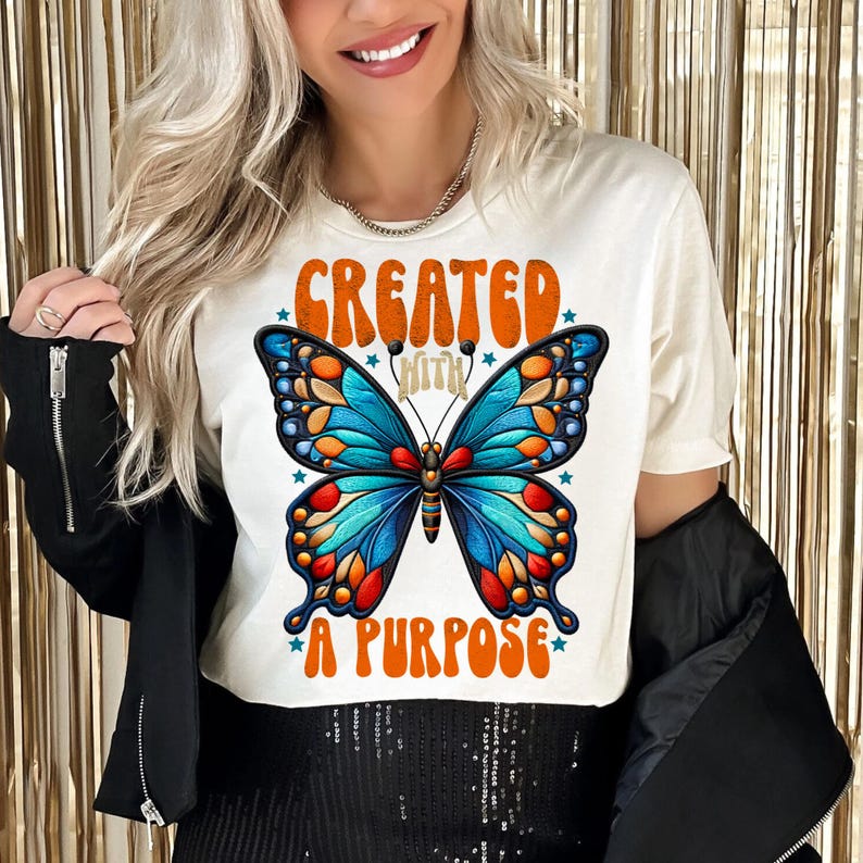 Pu&ograve; includere: Maglietta bianca con una grafica di farfalla colorata e il testo "Creato con uno scopo" in arancione.