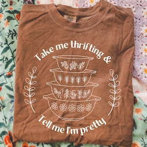 以下が含まれることがあります： 「Take me thrifting & Tell me I'm pretty」の白文字が入った茶色のTシャツ。花柄や鳥のデザインが施された重ねられたボウルのグラフィックが特徴です。Tシャツは折りたたまれています。