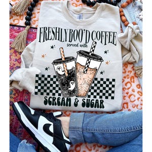 May include: A light beige sweatshirt with the text "Freshly Boo'd Coffee served with Scream & Sugar" and a graphic of two coffee cups with ghosts and spiderwebs.