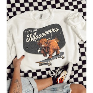 May include: White sweatshirt with a graphic of a brown and white highland cow skateboarding. The graphic is in a retro style with the text "I got the Mooooves".