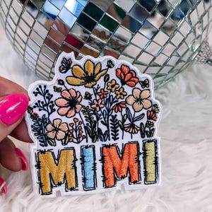 May include: An embroidered patch featuring a colourful floral arrangement above the word MIMI in block letters. The flowers are in shades of yellow, pink, and orange, while the letters are outlined in black with yellow, blue, orange, and green fill.