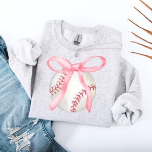 May include: A gray sweatshirt with a white baseball with a pink bow on it.