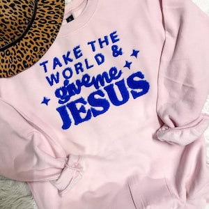 以下が含まれることがあります： 「TAKE THE WORLD & give me JESUS」と青い文字がプリントされた、薄いピンク色のスウェットシャツ。左上にヒョウ柄の帽子が見えます。スウェットシャツは長袖で、クルーネックです。