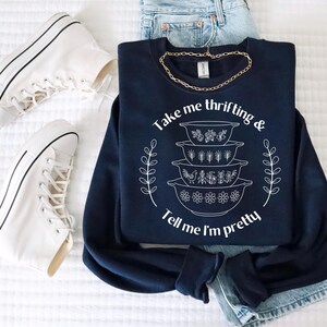 Op de afbeelding: Marineblauwe sweatshirt met de tekst "Take me thrifting & Tell me I'm pretty" en een afbeelding van gestapelde kommen. Witte high-top sneakers en een gouden ketting zijn ook zichtbaar.