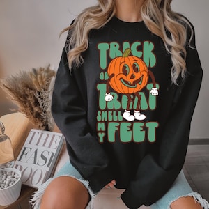 May include: Black sweatshirt with a retro-style Halloween design. The graphic features a smiling orange pumpkin with the text "Trick or Treat Smell My Feet" in green and white. A fun and festive apparel item for the spooky season.