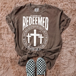 May include: A brown t-shirt with a white graphic design that says "REDEEMED" with a circle around it. The circle contains three crosses and the text "GALATIONS 2:20 I HAVE BEEN CRUCIFIED WITH CHRIST. AND IT IS NO LONGER I WHO LIVE, BUT CHRIST WHO LIVES IN ME."