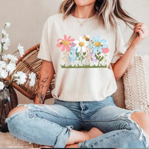 May include: Beige t-shirt featuring a colorful floral design with pink, white, and blue flowers, and a bee. The graphic is set against a green grass base. The shirt is a casual, comfortable style.