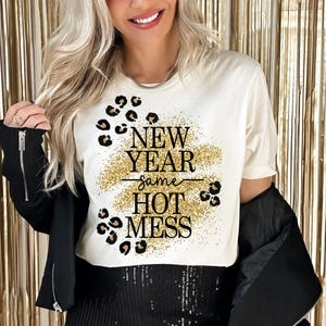 Può includere: Maglietta bianca con glitter dorati e stampa leopardata nera. Il testo sulla maglietta recita "New Year Same Hot Mess".