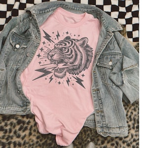 May include: Pink t-shirt with a black and white graphic of a tiger's head with lightning bolts and stars.